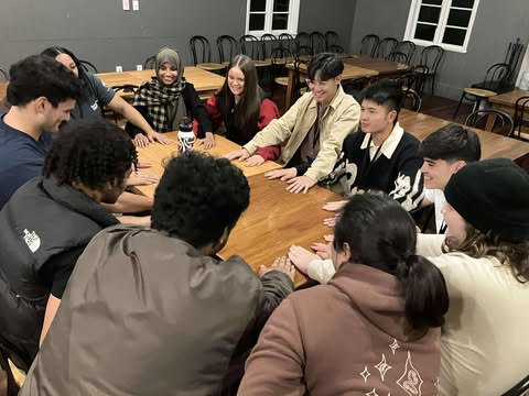 Young people at PALM 2025 are pictured sitting around a large wooden table. They are of various ethnicities and all have their hands stretched out in front of them. Many of them are smiling and looking up.