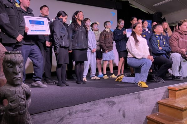 In this photo winners of the different categories in this year’s Ka Rere Te Manu Rangatahi Film Awards 2025 collect on stage. They fill the stage, with some kneeling and the others standing in a semi circle behind them. One teenage boy is holding a framed certificate. The young people range in age from pre-teens to teenagers.
