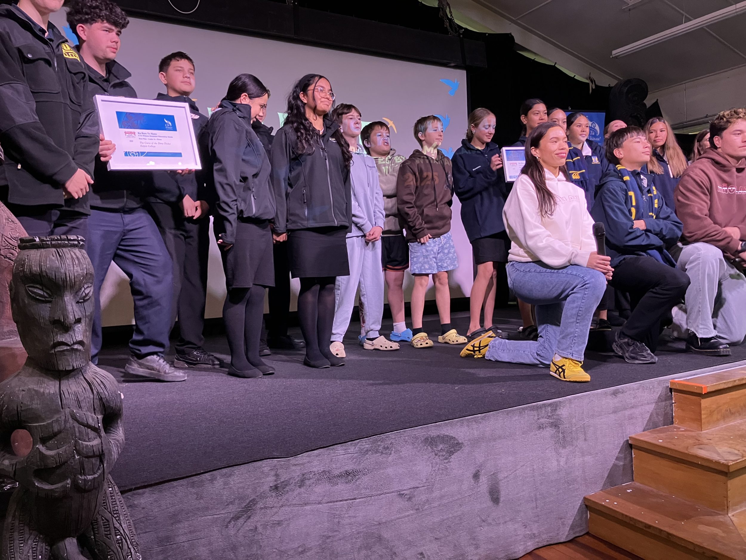 In this photo winners of the different categories in this year’s Ka Rere Te Manu Rangatahi Film Awards 2025 collect on stage. They fill the stage, with some kneeling and the others standing in a semi circle behind them. One teenage boy is holding a framed certificate. The young people range in age from pre-teens to teenagers.