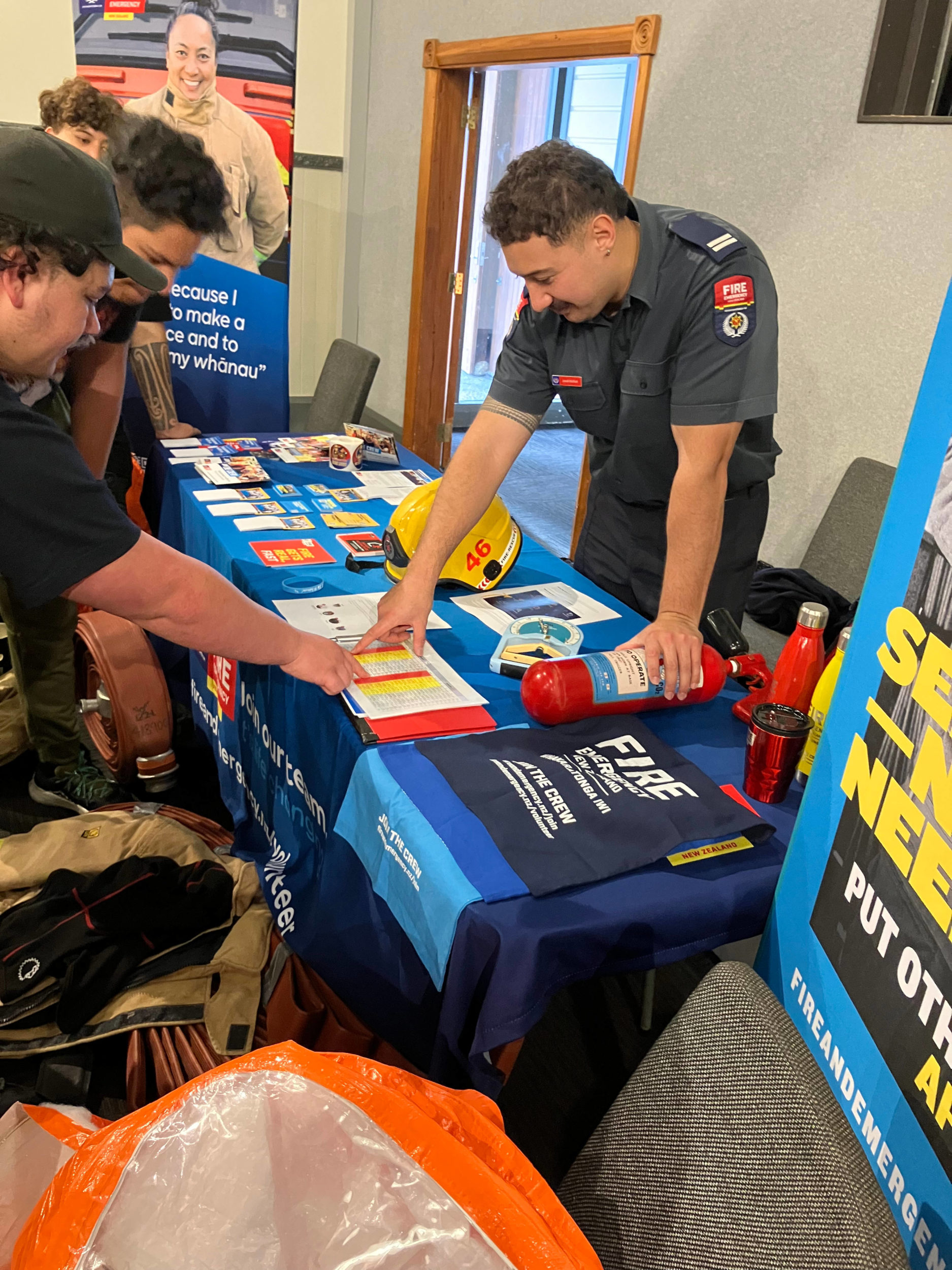 In this photo, a member of Fire and Emergency New Zealand (FENZ) hosts a stall at the careers expo. He is pointing at a document on a table. On the other side of the table, a small group of young men are looking on. Various FENZ documents, posters and other branded items are visible on the table and surrounding walls.