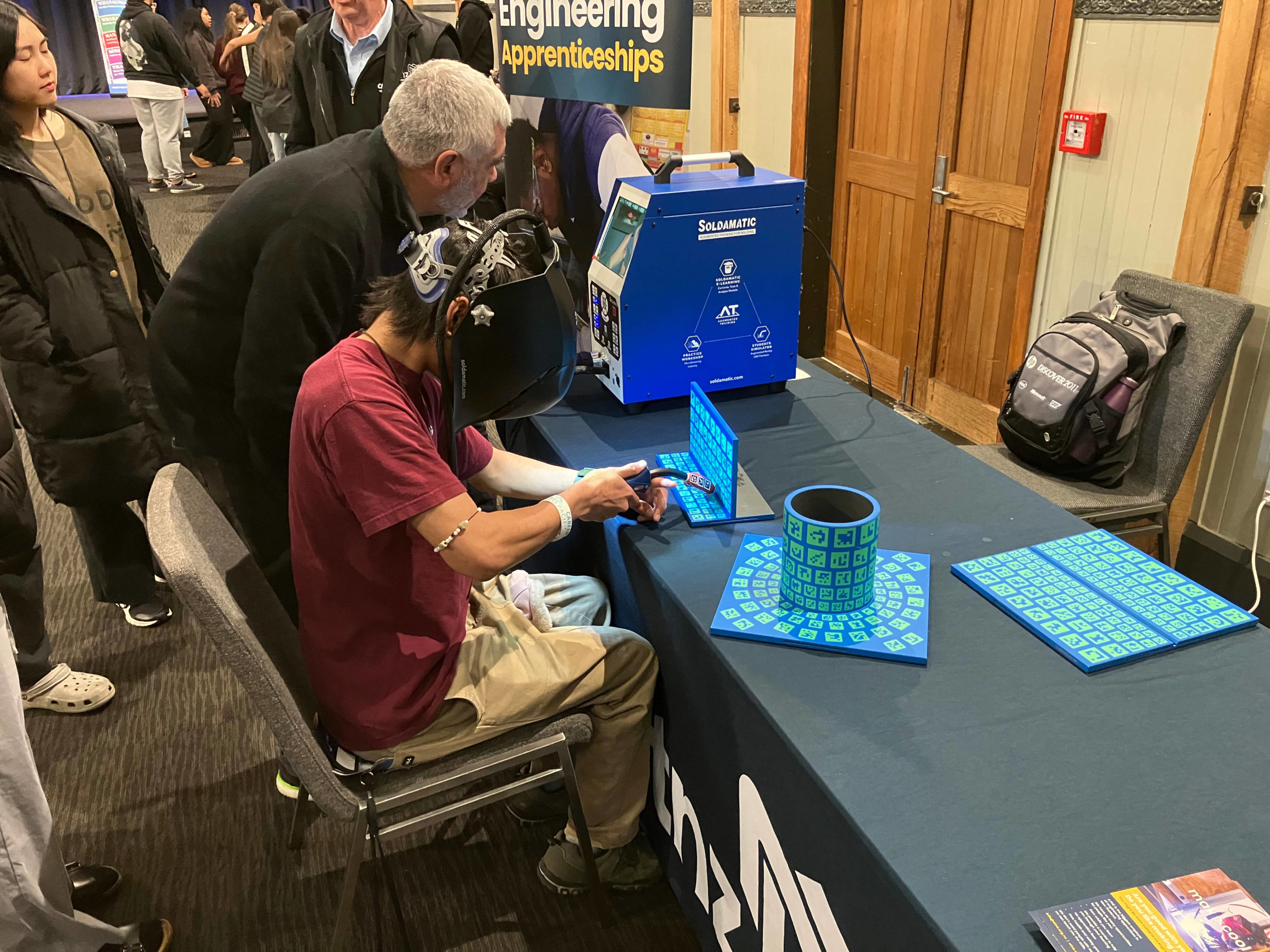 A young man tries a welding simulator. He is seated at at a table wearing a welding mask and holding a welding torch to a digital screen. The screen is linked to a welding simulator machine. An older man stands nearby and appears to be instructing the young man. Behind them, various other people are partially visible as they look on.