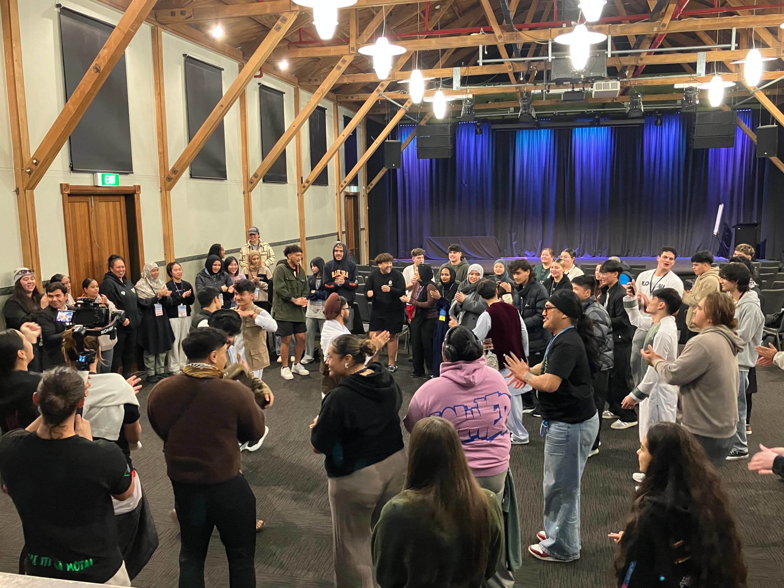 A large group of young people of various cultures and ethnicities stand in a circle at PALM 2025. They are all facing inwards, watching 5 young people who are dancing together. Many people appear to be clapping.