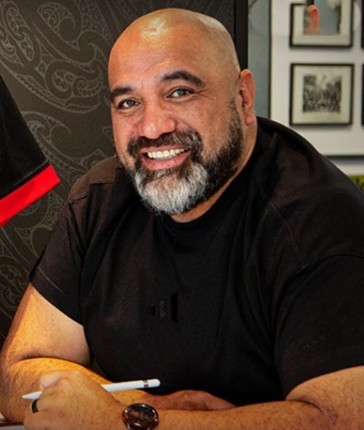 A head and shoulders shot of Kahurangi Falaoa, one of the judges in E Tū Whānau Poster Competition 2025. He is smiling at the camera and is holding a pen. Behind him various toi Māori art can be seen.