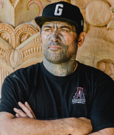 A head and shoulders shot of Stu McDonald, one of the judges in E Tū Whānau Poster Competition 2025. His face is patterned with tā moko and he wears a black cap and t shirt.