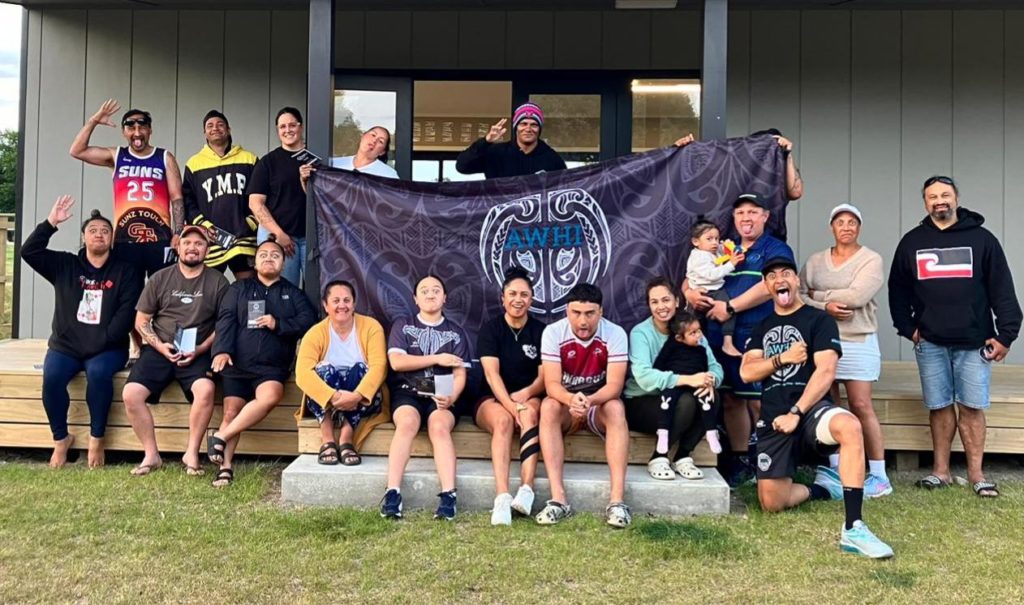 Young AWHI influencers are pictured on the step of a building. About twenty people either sit or stand around a large blue and black flag in the centre of which is the AWHI logo.