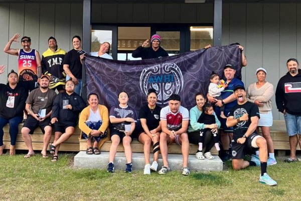 Young AWHI influencers are pictured on the step of a building. About twenty people either sit or stand around a large blue and black flag in the centre of which is the AWHI logo.