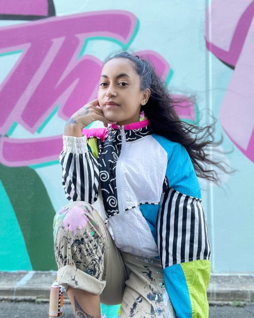 A head and shoulders shot of Miriama Grace-Smith one of the judges in E Tū Whānau Poster Competition 2025. She wears bright coloured and multi patterned clothing and is looking at the camera. Behind her is part of brightly coloured wall mural