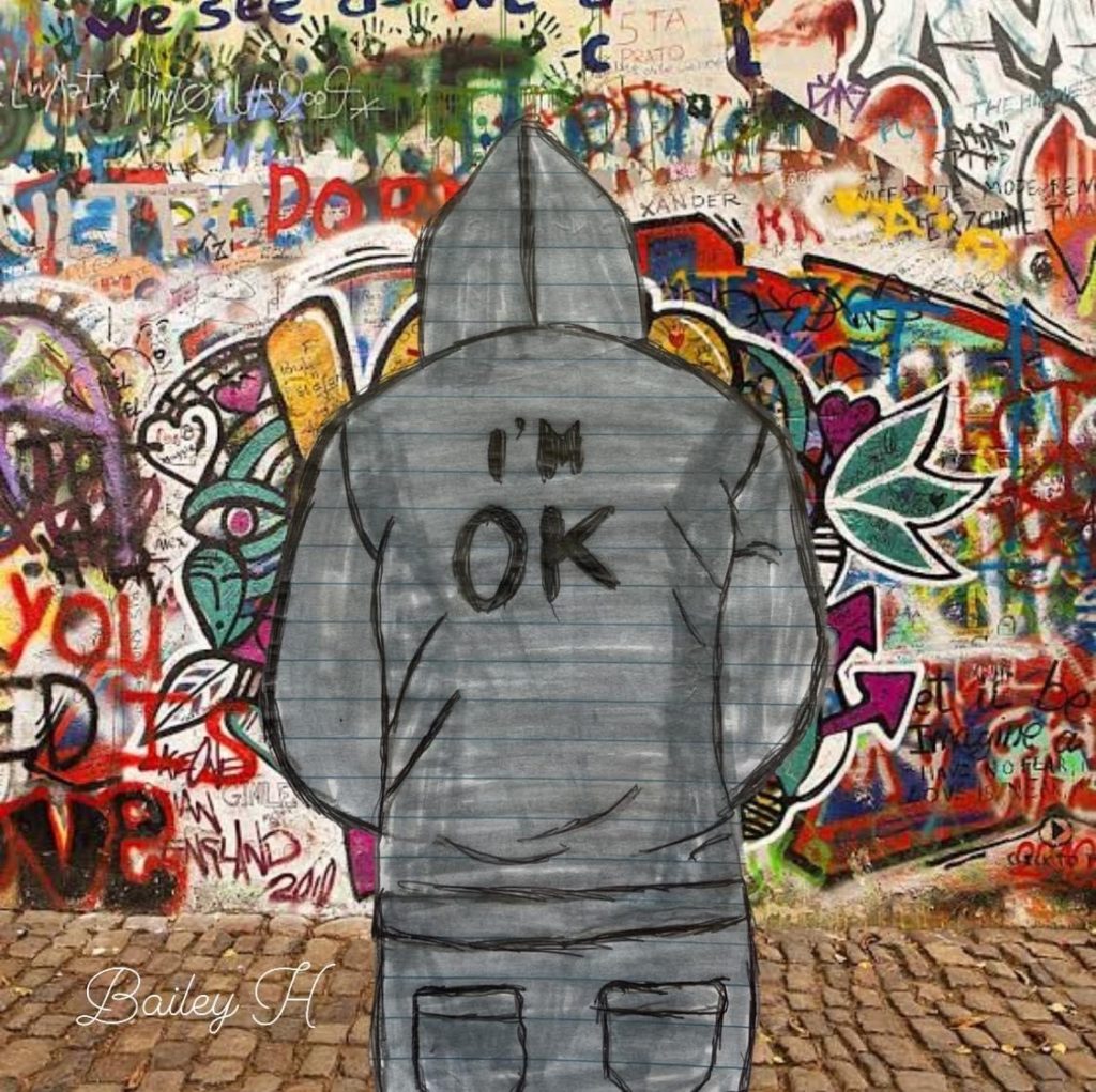 An image of Bailey Harrison's Highly Commended entry in the 13 to 18 years category of the poster competition - one of the community events run by E Tū Whānau in 2025. It depicts a male figure who is looking away form the viewer and wearing a grey hoodie, the back of which says "I'm ok". The figure is in front of a a heavily graffitied wall. 