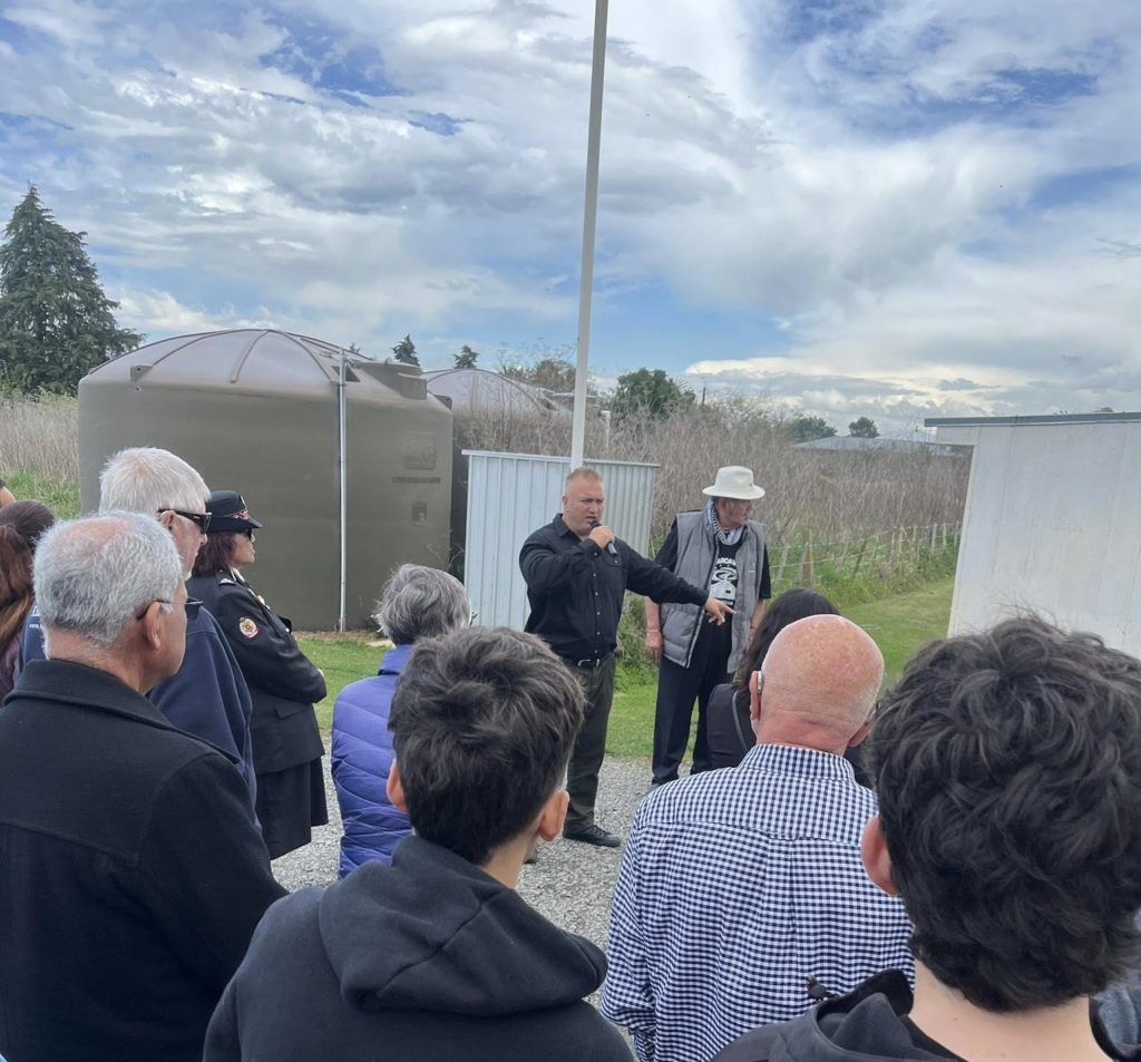 Thompson Hokianga is pictured standing in the grounds of a marae at one of the open days speaking to a group of people. While they are all faced away from the camera, Thompson is seen holding a microphone as he speaks. He is dressed in dark clothing.