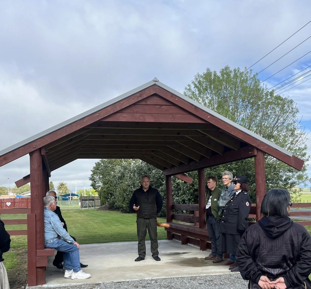 Thompson Hokianga is pictured standing in the mahau, or covered entry way, to a marae on one of the open days. He is middle-aged, dressed in dark clothing and appears to be speaking to various people who are gathered around. Some are seated, some are standing.