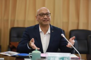 Toa Faneva, Chairperson of Māori Reference Group, is pictured in business attire. He is seated before a microphone. He gestures with his hands and appears to be talking.