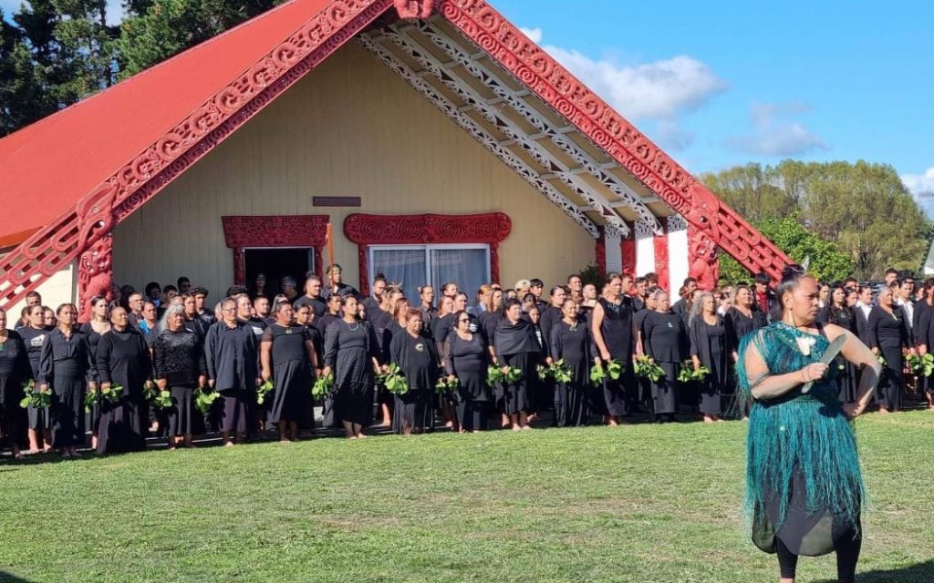 TThis photo shows a woman wearing a green dress and holding a mere pounamu as she calls the karanga at the pōwhiri at Ōmahu Marae on one of the open days. Behind her, many whānau stand in lines. they are dressed in black, holding green leaves and stand in front of the ornately carved maihi beams on the front of the whare nui.