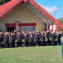 TThis photo shows a woman wearing a green dress and holding a mere pounamu as she calls the karanga at the pōwhiri at Ōmahu Marae on one of the open days. Behind her, many whānau stand in lines. they are dressed in black, holding green leaves and stand in front of the ornately carved maihi beams on the front of the whare nui.