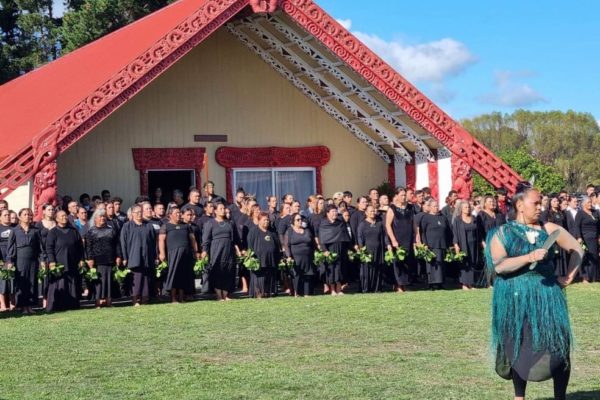 TThis photo shows a woman wearing a green dress and holding a mere pounamu as she calls the karanga at the pōwhiri at Ōmahu Marae on one of the open days. Behind her, many whānau stand in lines. they are dressed in black, holding green leaves and stand in front of the ornately carved maihi beams on the front of the whare nui.