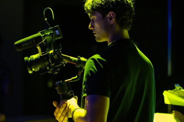 Isaac Te Reina is pictured while filmmaking from behind. the light is moody and he is holding a camera.