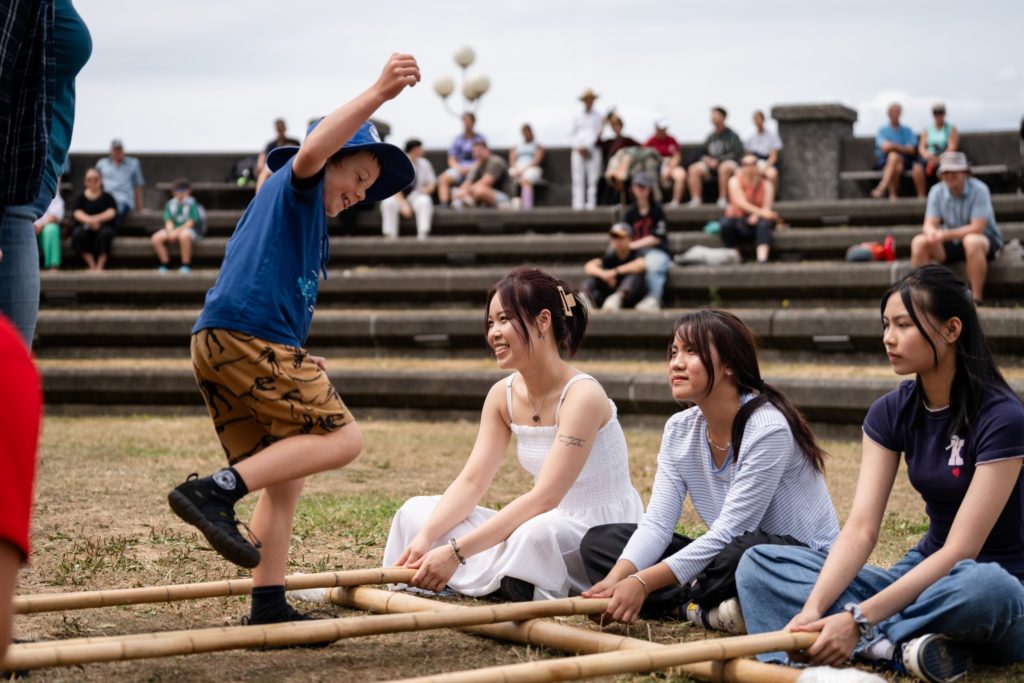 A child steps between two sets of bamboo poles on grass while three seated young women hold and move the poles, teaching the child the traditional Vietnamese bamboo dance called múa sạp. This cultural activity is often performed during festivals such as Lunar New Year. People are sitting on tiered stone steps in the background watching the activity.