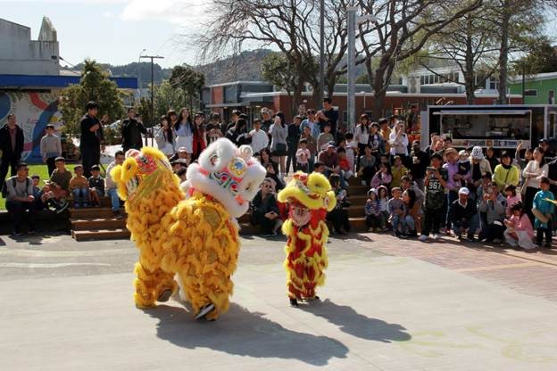 Three Viet Hub NZ performers in bright yellow lion costumes with white and multicolored lion heads dance in an outdoor courtyard during the Vietnamese Trung Thu (Mid-Autumn) Festival in Wellington. A large crowd of seated and standing spectators watches in the background, with trees, buildings, and a food truck visible under clear daylight.