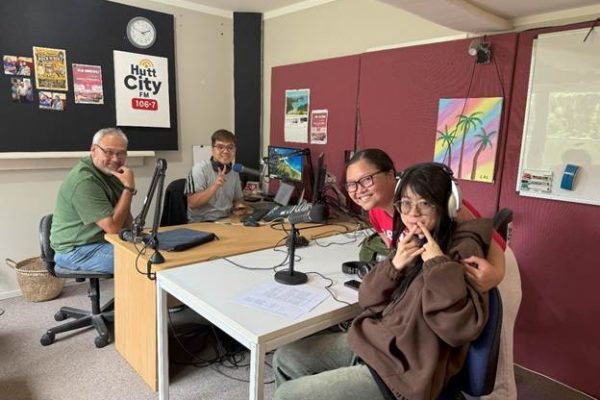 Three individuals from Viet Hub NZ sit at a table in a radio studio during their first trial broadcast with Hutt City FM 106.7. Microphones, headphones, and audio equipment are visible on the desk, and a large window provides natural light in the background.