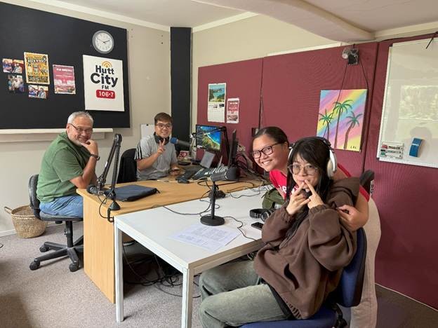 Three individuals from Viet Hub NZ sit at a table in a radio studio during their first trial broadcast with Hutt City FM 106.7. Microphones, headphones, and audio equipment are visible on the desk, and a large window provides natural light in the background.