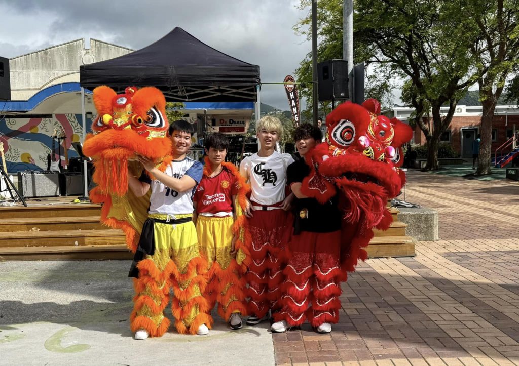 Four Viet Hub NZ performers standing outdoors holding two vibrant lion dance costumes with orange and red fur and large decorative heads, during a Vietnamese street festival. A stage with speakers and a mural is visible in the background.