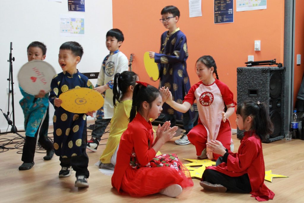 In this photo, eight young Vietnamese children, many wearing cultural dress, joyfully performing a song to celebrate the Mid-autumn Festival 2024 at Te Mako Centre, Naenae. Four young girls are seated and appear to be clapping each others' hands while the boys dance in a circle behind them.