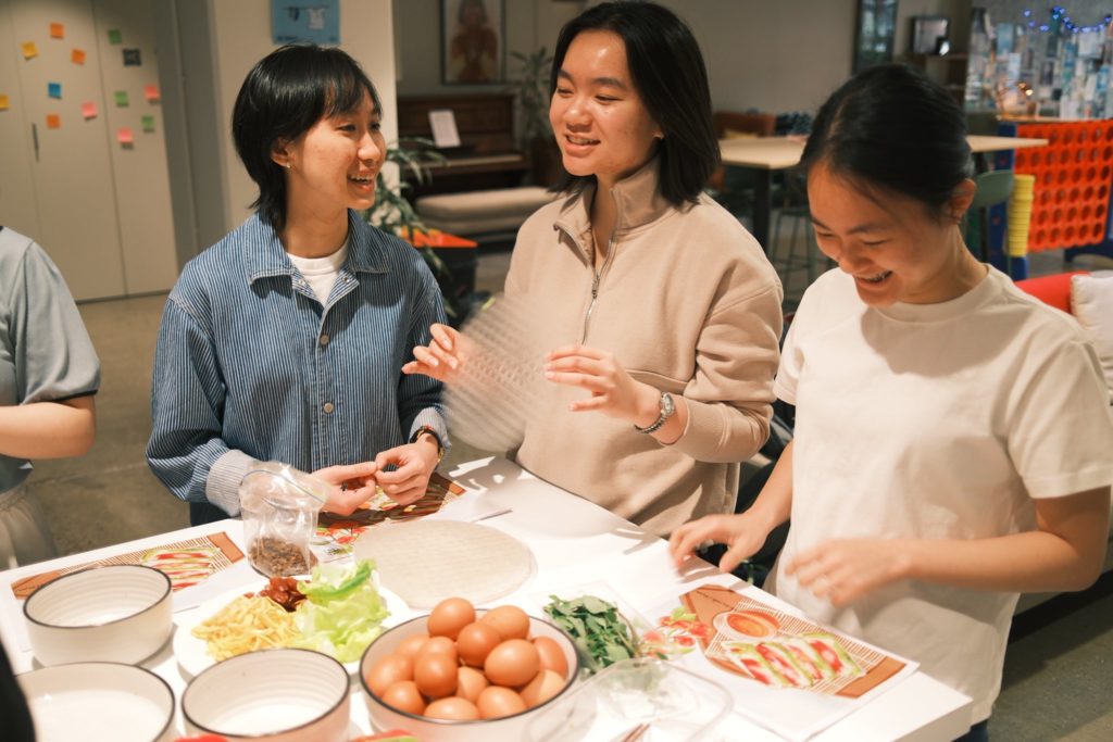 Three young Vietnamese people chat while cooking at a Viet Hub NZ organised cooking class. They look happy and appear to be following a recipe. On the table in front of them are eggs, sliced green vegetables and empty bowls.