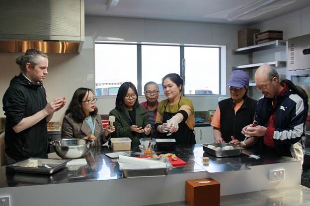 Participants gather around a kitchen counter during a Vietnamese cooking workshop organised by Viet Hub NZ, learning how to make traditional mooncakes. Ingredients, mixing bowls, and trays are arranged on the counter.