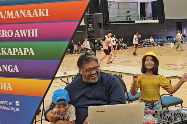 In this photograph taken at Poitūkohu Māori National Championships 2026, an older man sits holding a young boy. Next to them is a young girl, playfully looking at the camera. In the foreground, is a flag showing the six E Tū Whānau values. They are inside an indoor sports venue.
