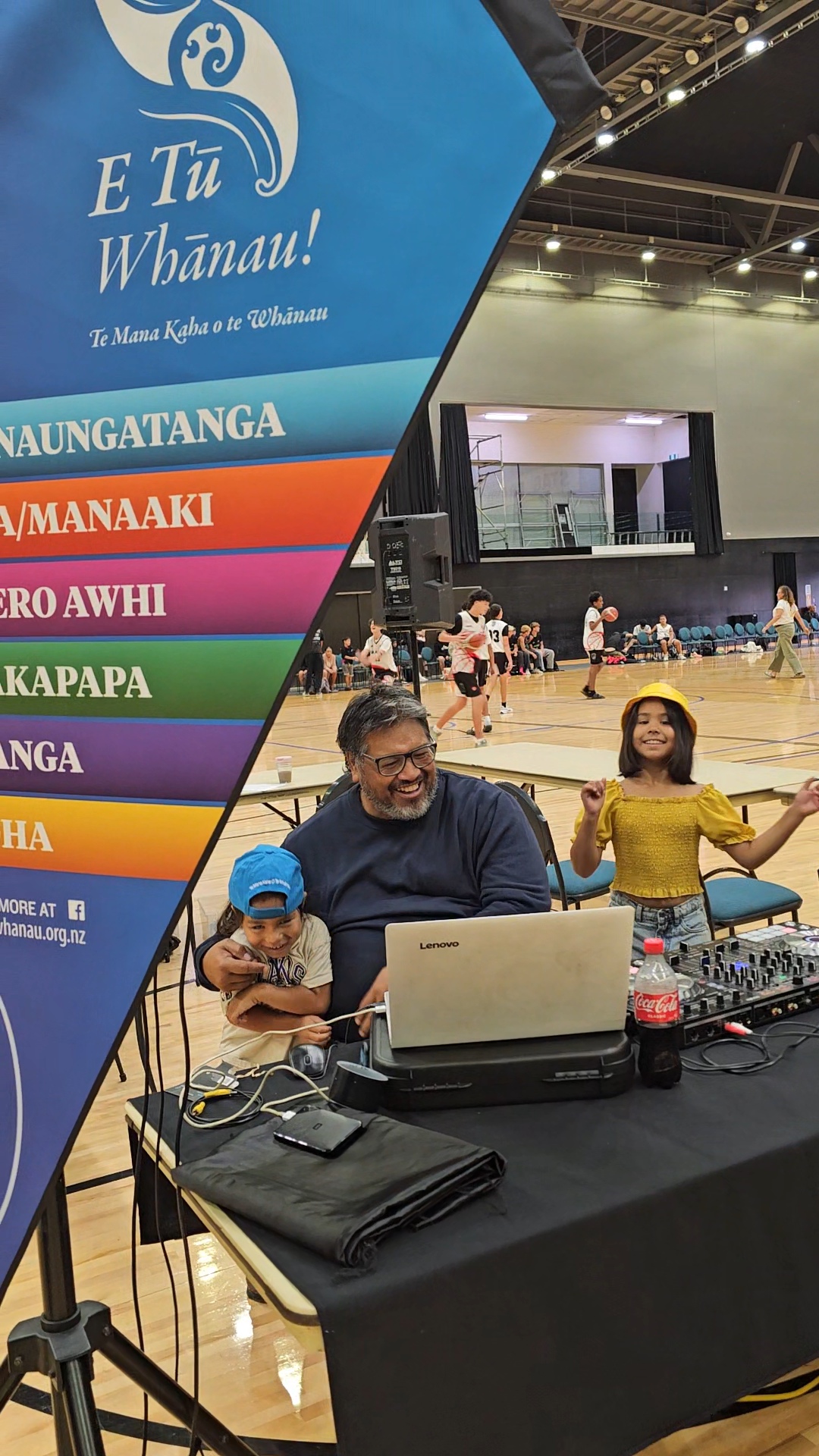 An older man sits holding a young boy. Next to them is a young girl, playfully looking at the camera. In the foreground, is a flag showing the six E Tū Whānau values. They are inside an indoor sports venue.