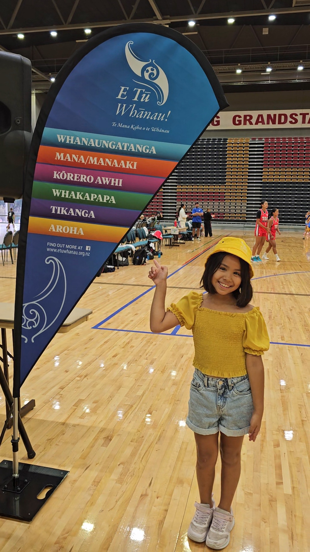 In this photograph taken at Poitūkohu Māori National Championships 202, a young girl happy points at an indoor flag of the six E Tū Whānau values. She is pictured inside an indoor sports venue.