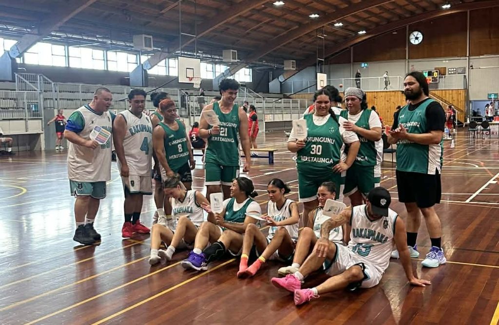 Heretaunga and Rakaipaaka teams are pictured together at Poitūkohu Māori National Championships 2026. Some are seated on the floor of the indoor court, while others stand behind. They all look sweaty and are holding E Tū Whānau values fans. 