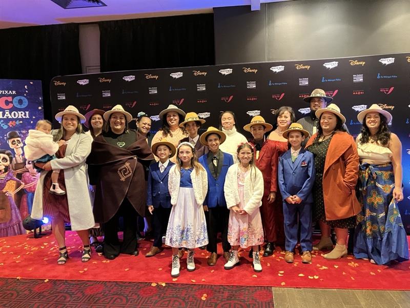 The Mānihera whānau and Adele Harrison smartly dressed. ON a board behind them, the words "Disney Pixar Coco Reo Māori" are partially visible. They look happy and are smiling.