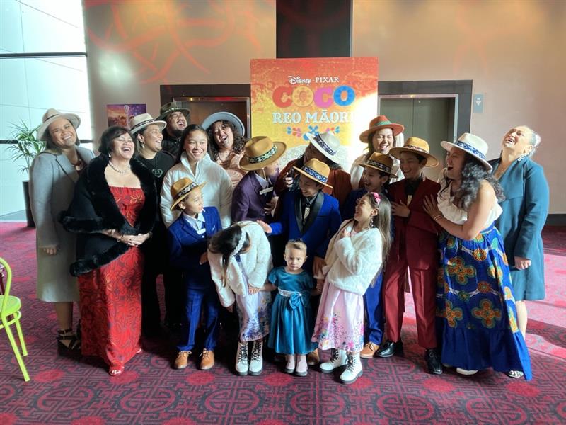 The Mānihera whānau and Adele Harrison smartly dressed and collected in a theatre foyer around a colourful pull up sign on which the words "Disney Pixar Coco Reo Māori" are visible. They look happy and many are laughing.