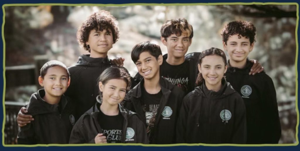 Seven children from the Mānihera whānau stand close together, some with arms about each other's shoulders, for a photo. They are aged between about 8 and 12 and are all smiling.