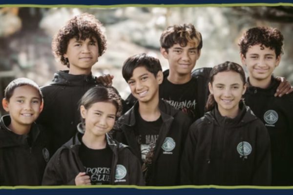 Seven children from the Mānihera whānau stand close together, some with arms about each other's shoulders, for a photo. They are aged between about 8 and 12 and are all smiling.