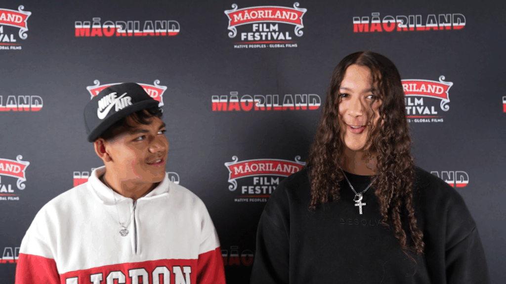 Two teenage boys are pictured. One wears a black cap and a red and white sweatshirt. He is looking at the other boy who is dressed in black and has shoulder-length hair