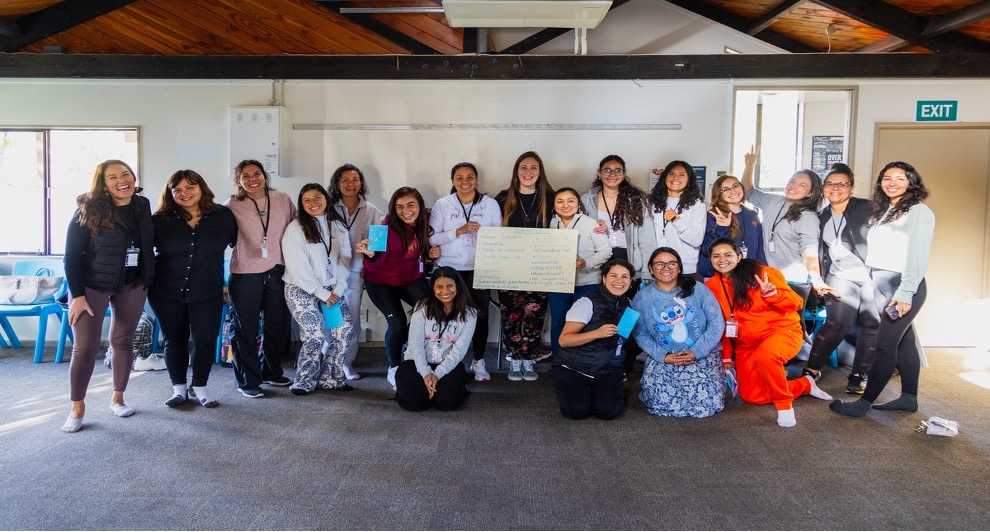 A large group of women stand and kneel together at an ALAC workshop. They are generally young and all look happy. The women in the middle hold a large piece of paper with indistinct writing on it