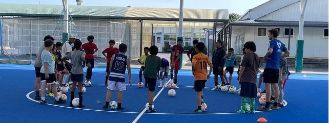 A group of young children stand in a circle on an indoor football pitch. Each has a football at their feet. One adult man is talking to them.
