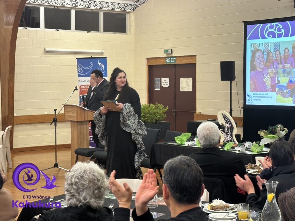 Candis Brunning stands in front of crowd of people while being recognised as a Rangatahi Kahukura – Emerging Leader at E Tū Whānau Kahukura Awards 2026. She is dressed in black and holds a certificate.
