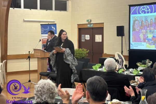 Candis Brunning stands in front of crowd of people while being recognised as a Rangatahi Kahukura – Emerging Leader at E Tū Whānau Kahukura Awards 2026. She is dressed in black and holds a certificate.