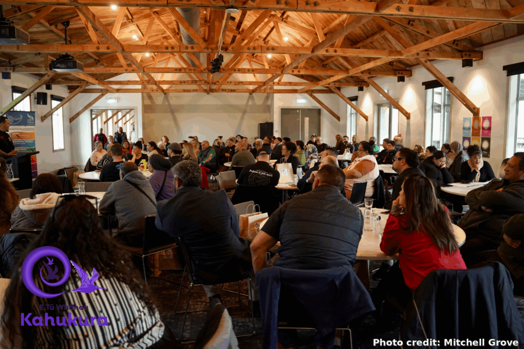 People are pictured sitting around large tables in a room at Kahukura Workshop 2026. The room is full - there appear to be over 100 people. All are paying attention to a speaker who is presenting at the front of the room.