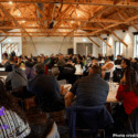 People are pictured sitting around large tables in a room at Kahukura Workshop 2026. The room is full - there appear to be over 100 people. All are paying attention to a speaker who is presenting at the front of the room.