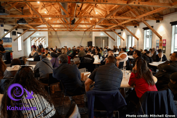 People are pictured sitting around large tables in a room at Kahukura Workshop 2026. The room is full - there appear to be over 100 people. All are paying attention to a speaker who is presenting at the front of the room.
