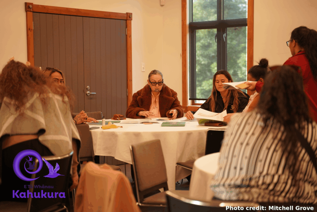 People are pictured in of the smaller break out groups at Kahukura Workshop 2026. Eight people - a mixture of men and women - sit around a large table. They are looking at documents, while a woman, whose back is to the camera, facilitates the session.