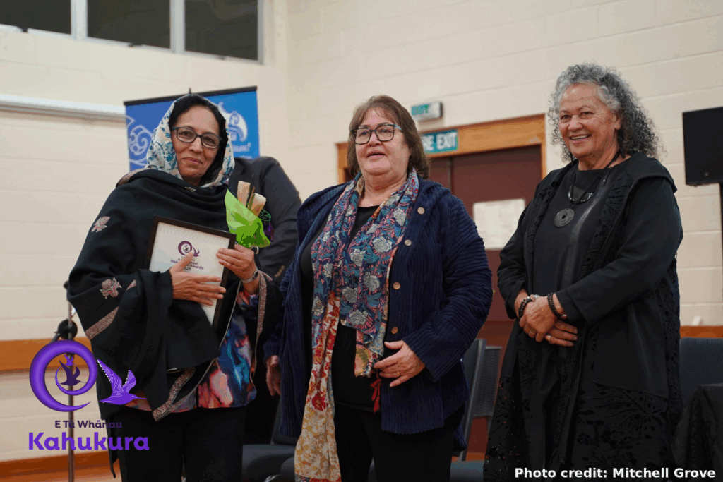 Fahima Saied, New Settlers Family and Community Trust CEO, is pictured wearing a headscarf and holding a Kahukura Awards certificate and flowers. She is with Heni Turner, E Tū Whānau Kaiwhakahaere, and Katie Murray. All three women are smiling.