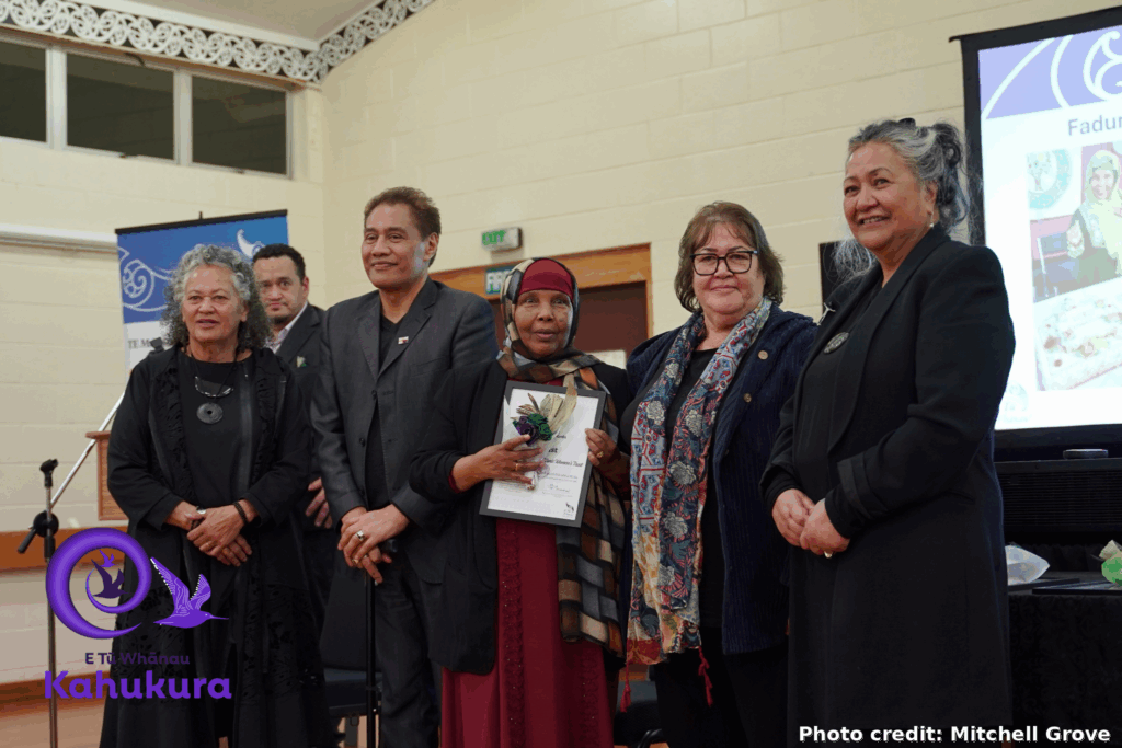 Fadumo Ahmed, QSO, is pictured wearing a head scarf and formal clothing while holding her certificate. She stands in a line with Katie Murray, Brent Mio, Darrin Haimona, E Tū Whānai Kaiwhakahaere Heni Turner, and Donna Matahaere-Atariki.