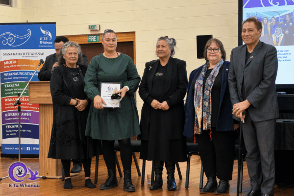 Renee Davis is pictured in a formal green dress at teh Kahukura Awards 2026 ceremony. She is holding a certificate and flowers and stands in a line with Katie Murray, Donna Matahaere-Atariki, E Tū Whānau Kaiwhakahaere Heni Turner, and Darrin Haimona.