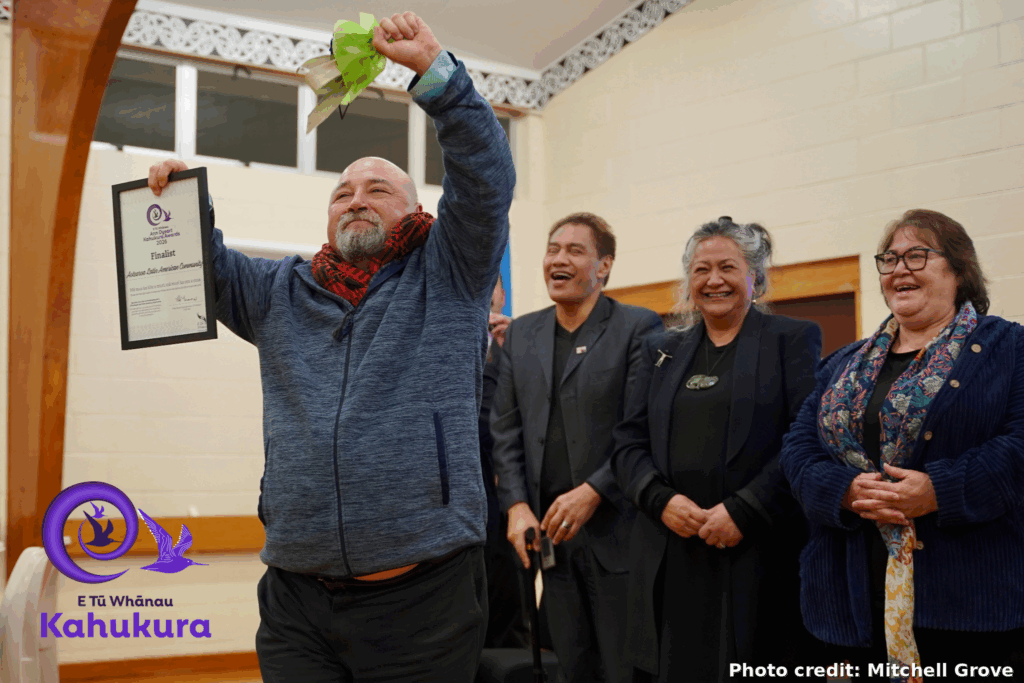 Esteban Espinoza, ALAC Founder and Director, celebrates ALAC being recognised as a finalist in the E Tū Whānau Ann Dysart Kahukura Award 2026. He has his hands raised in the air and is smiling. He is holding his finalist certificate and flowers. Behind him, people can be seen smiling and laughing.