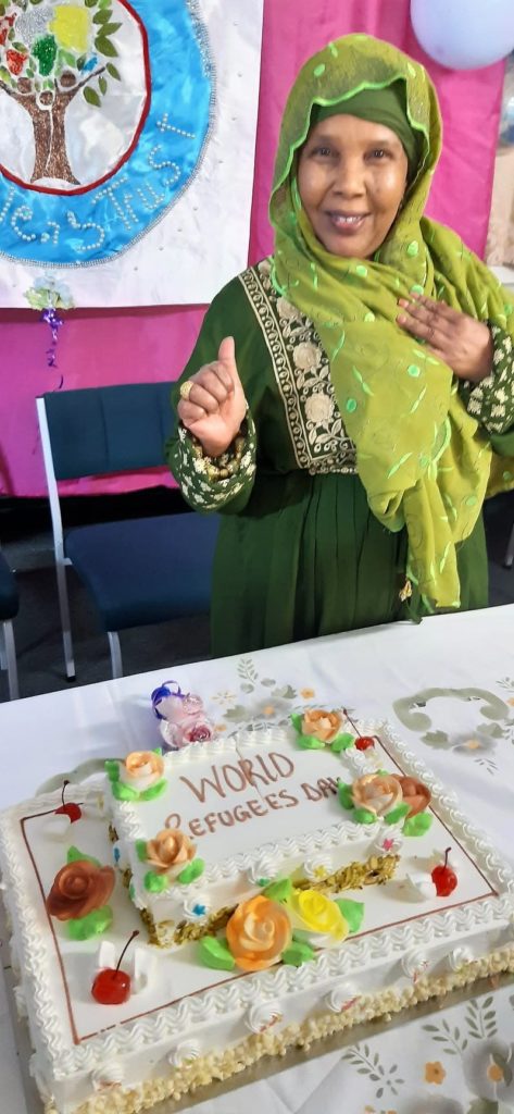 Fadumo Ahmed, dressed in a green dress and headscarf, stands before a colourful decorated cake that has "World Refugees Day" written on it.