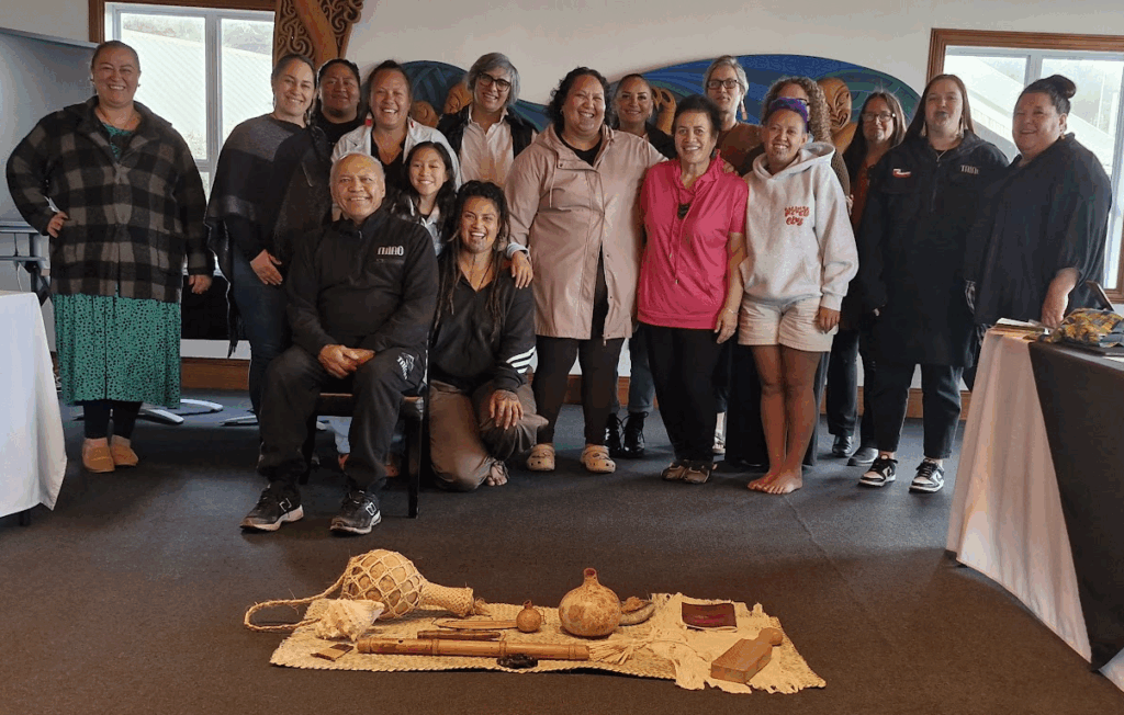 A group of about 15 people connected to Mana Wāhine Collective stand together in doors. Some are standing and some are seated or kneeling. On the ground in front of them are some wooden taonga on a woven flax mat.